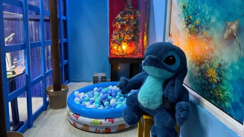 Velika plišana igračka Stitch na stolici, pored bazenčića sa lopticama i vitrine sa ukrasima.