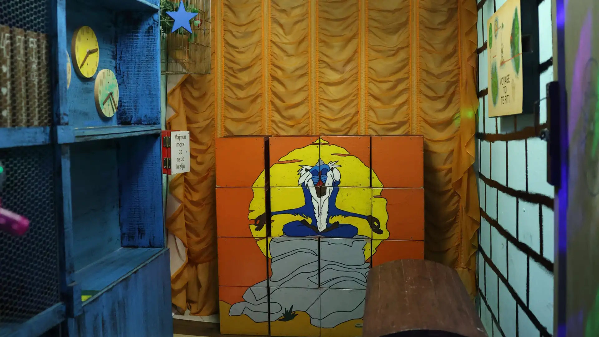 Unutrašnjost Bajka escape room sobe sa plavim drvenim policama, dva zidna sata, zlatnim zavesama i slagalicom sa likom Rafikija iz Kralja lavova.
