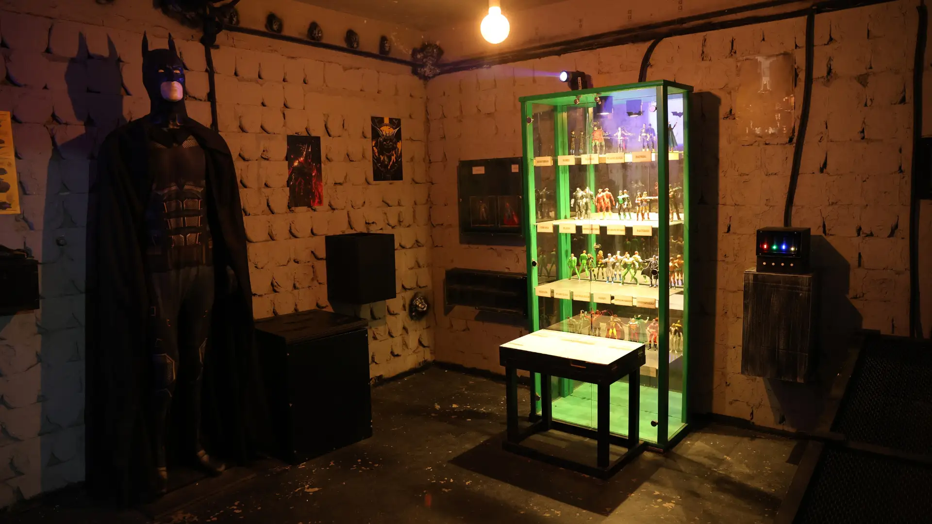 Betmen u crnom kostimu pored osvetljene vitrine sa figuricama superheroja u Betmen escape room sobi.