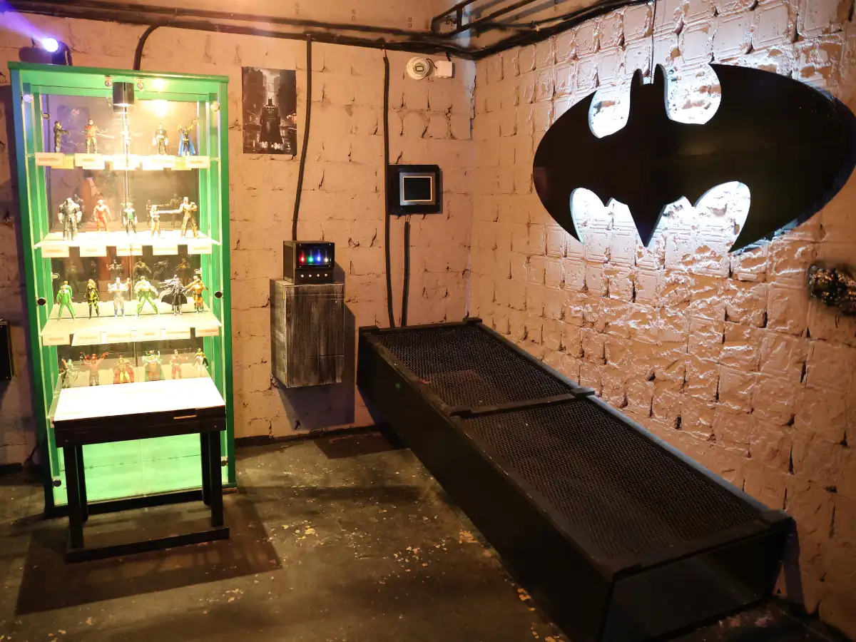 Betmen escape room sa osvetljenim znakom šišmiša na zidu i vitrinom sa figurama superheroja
