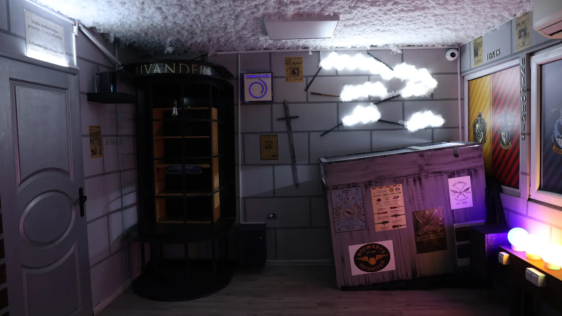 Unutrašnjost Hari Poter escape room sobe sa vratima, policom sa knjigama, Hogwarts sandukom, grbovima čarobnjačkih kuća i dekorativnim oblacima koji svetle.
