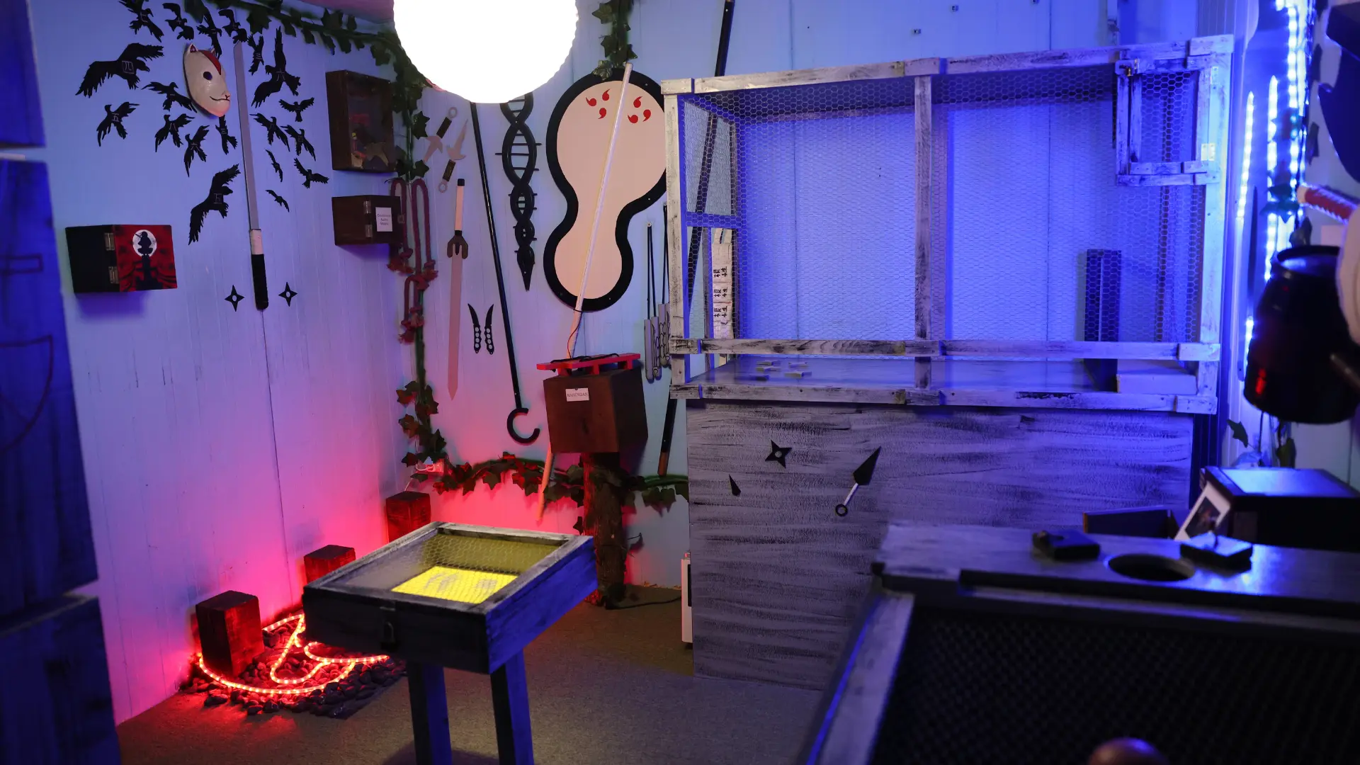 Unutrašnjost Naruto escape room sobe sa nindža oružjem, šurikenima, kunai noževima i zagonetnim rekvizitima pod plavim i crvenim osvetljenjem.