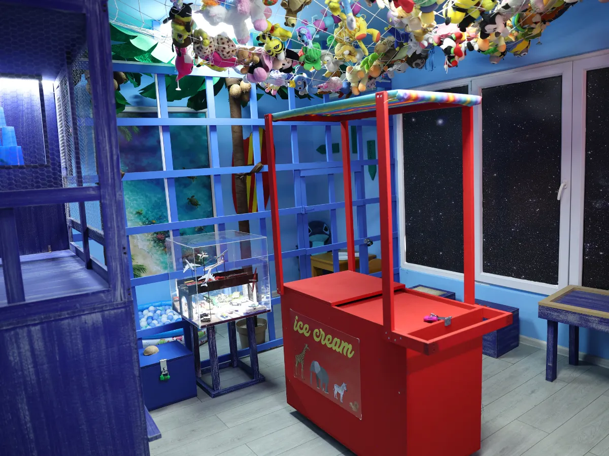 Unutrašnjost escape room sobe Stič sa crvenim štandom za sladoled, plišanim igračkama na plafonu i plavim dekoracijama.