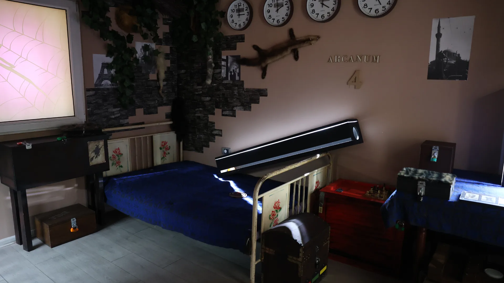 Unutrašnjost Veštica escape room sobe sa krevetom prekrivenim plavim prekrivačem, drvenim sanducima, četiri zidna sata i osvetljenim panelom sa motivom paukove mreže.